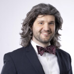Smiling man with long gray hair in suit and patterned bow tie, prepared for Apologetics Day against a plain background.
