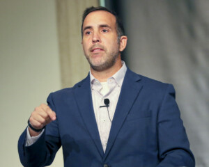 Speaker in a blue blazer gestures during Apologetics Day, using a microphone headset at an academic event indoors.