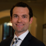 Smiling man in suit and tie at Apologetics Day indoors, soft arches and warm lights blurred in the academic event background.
