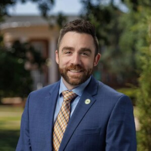 Man in blue suit with plaid tie smiling outdoors on HCU campus, trees and building in background, likely at a Pinson Lecture event.