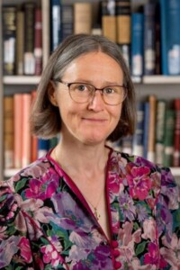 Woman with glasses and short brown hair in floral blouse stands by bookshelves after A.O. Collins academic lecture.