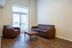 Cozy campus lounge with brown sofa, armchair, and wooden coffee table by large windows letting in natural light.
