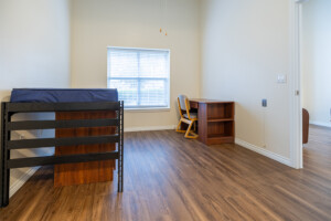 University Place dorm room featuring a bed, wooden desk, chair, and window with blinds on hardwood flooring.