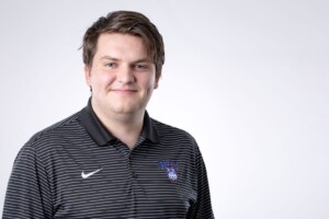 Smiling man with brown hair in a black striped polo stands before a white backdrop, representing Residence Life at Houston Christian University.