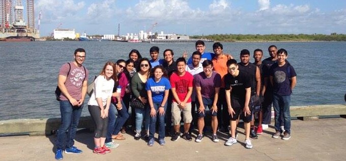 Group stands on a sunny HCU campus dock by the water at an event, with industrial buildings in the background.