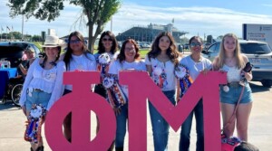 Seven women with a dog stand by pink ΦΜ letters on HCU campus, all wearing colorful homecoming mums outdoors.