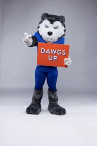 Kiza, the HCU husky mascot, holds an orange DAWGS UP sign and flashes a peace sign on HCU campus during a spirited event.