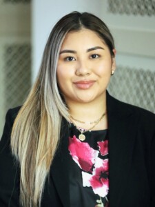 Smiling woman with long blonde hair in a black blazer and floral top, indoors, possibly in an academic or professional setting.
