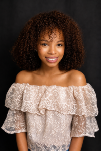 Smiling woman with curly hair in a lacy white top, SHRM member, standing against a dark backdrop on HCU campus.