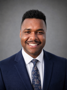 Smiling man in a blue suit and floral tie poses, reflecting Houston Christian University professionalism, on gray backdrop.