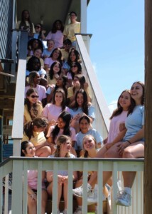 Smiling young women in pastel shirts on stairs outside a wooden building on HCU campus, representing Alpha Chi Omega.