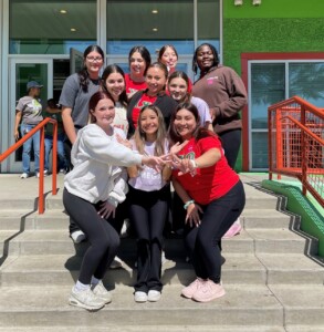 Ten Alpha Chi Omega members smile on HCU campus steps outside a modern building with large glass doors.