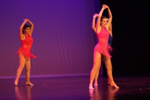Dance students in red costumes perform on stage with arms raised, purple-lit backdrop in Dunham Theater.