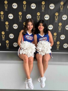 Two dance squad cheerleaders smile with white pom-poms, seated indoors before a trophy backdrop at a university event.