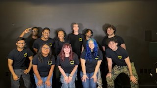 Improv Club members in matching black shirts smile together during a drama workshop against a plain dark backdrop.