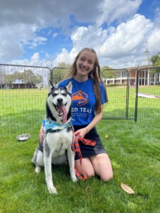 Woman kneels on grass beside happy Husky, Kiza, wearing blue bandana outside HCU campus building.