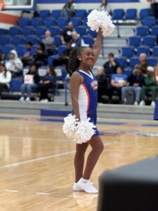 Cheerleader in blue and white uniform smiles, holding pom-poms on basketball court with spectators in background.