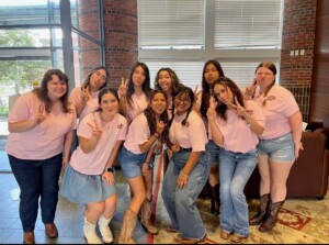 Group of ten women in pink and denim pose indoors, smiling, making peace signs; several wear cowboy boots.