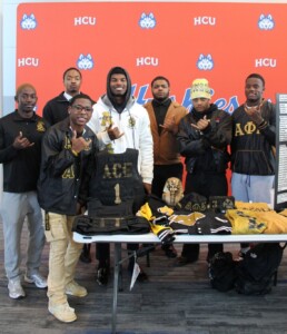 Eight Alpha Phi Alpha members stand with fraternity items in front of an HCU backdrop on the Houston Christian campus.