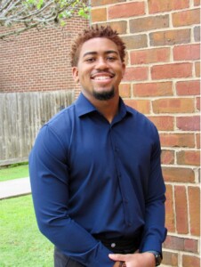Student government member in a blue shirt smiles by a brick wall and fence on HCU campus on a sunny day.