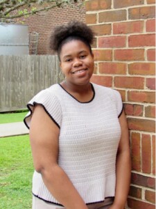 Student Council member smiles, leaning on HCU campus brick wall, with grass and fence in the background.