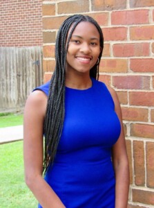 Smiling woman in a blue dress with long braids stands for student leadership outside brick wall on HCU campus.