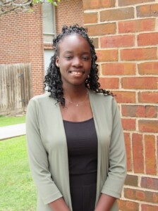 Student Government member with curly hair in a green jacket, smiling near a brick wall on HCU campus.