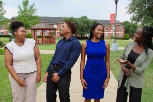 Four students laugh together on an HCU campus sidewalk; Student Center and campus organizations visible in the background.