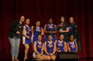 Husky Dance Squad members in blue and casual wear pose with flowers onstage before a red theater curtain.