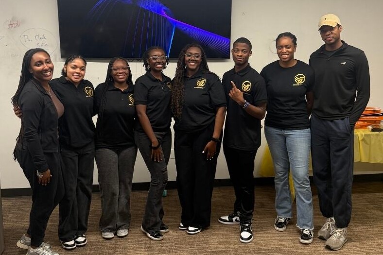 Eight people in black attire smile together in an office, gathered around a table and TV, celebrating their achievement.