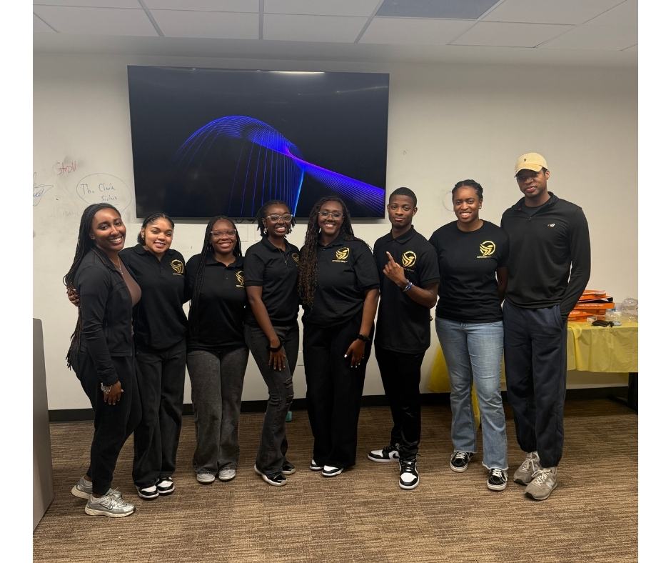 Eight people in black attire smile together in an office, gathered around a table and TV, celebrating their achievement.