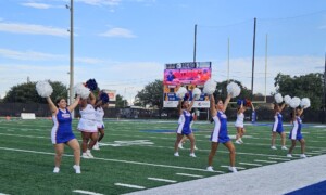 HCU Dance Squad in blue uniforms performs with pom-poms on the football field under stadium lights.