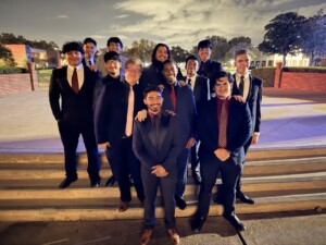 Thirteen Phi Kappa Psi members in suits smile on HCU campus stairs under bright lights at night.