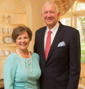 Smiling woman in a light blue dress stands with a man in a dark suit and red tie at an indoor academic event under warm lighting.