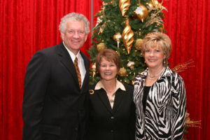 Three adults in formal attire smile beside a Christmas tree, sharing memories during an HCU campus holiday alumni gathering.