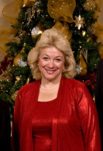 Evelyn Husband Thompson in a red dress smiles by a Christmas tree, enjoying memories from previous Christmas Luncheons during campus festivities.