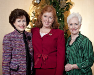 Three formally dressed women smile at a Christmas luncheon beside a decorated tree with gold ribbon, attending an academic event.