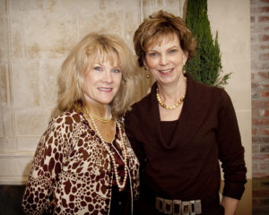 Two women smiling at a Guild Fall Coffees event, standing by a stone wall with greenery, indoors at a campus gathering.