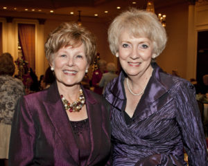 Two older women in purple outfits share a smile at an indoor Christmas event, celebrating with fellow attendees at a university gathering.