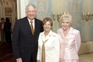 Three formally dressed older adults smile together at a Silver Teas event in an ornate room with patterned wallpaper.