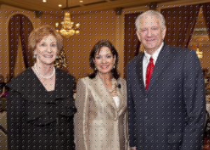 Three adults in formal wear smile at a festive luncheon beneath chandeliers and curtains, enjoying a university holiday gathering.