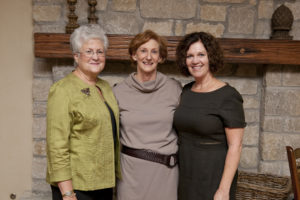 Three women smiling together indoors by a stone fireplace mantel during a university event, possibly in an academic building lounge.