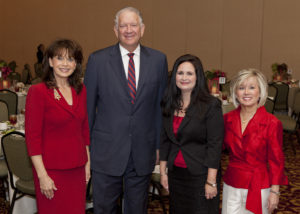 Professionals in business attire smiling at a holiday luncheon, seated around round tables inside an academic event space.