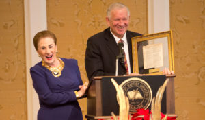 A man smiles, holding a framed document at a podium during a festive luncheon, laughter shared among attendees indoors.