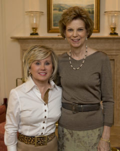 Two women at a Guild event, smiling together by a fireplace indoors, both wearing neutral attire in an academic setting.