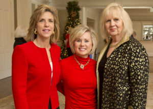 Three women share smiles beside a decorated Christmas tree during a festive campus luncheon, celebrating the holiday season.