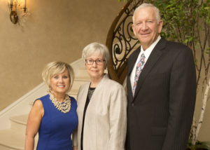 Three formally dressed older adults standing together at a Silver Teas event by a staircase and ornate railing indoors.