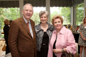 Three older adults smiling together at a Silver Teas event indoors, with large windows and lush greenery in the background.