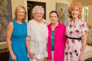 Four smiling women in colorful dresses stand indoors, with artwork and light walls suggesting an academic or gallery setting.