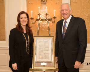 A woman and man in formal wear stand by a framed document during a festive academic luncheon in an elegant indoor setting.